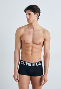 Juodi medvilniniai bokseriai su balta "CALVIN KLEIN" logotipu ant juosmens, siūlantys prigludusį dizainą ir lygią tekstūrą. Modelis be viršutinės kūno dalies.
