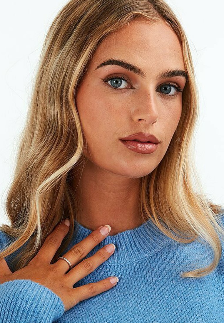 Blauer gestrickter Pullover mit einem strukturierten Muster und rundem Ausschnitt. Das Modell trägt einen schlichten silbernen Ring an der linken Hand.