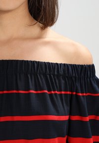 Top off-the-shoulder in tessuto blu navy con righe orizzontali rosse, con scollatura elastica e texture morbida.