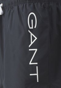 Juodos maudymosi šortai, pagaminti iš lengvo audinio, turi elastingą juosmenį su raišteliu ir didelį baltą "GANT" logotipą šone.