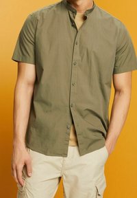 Man in olijfgroen overhemd met korte mouwen over een beige T-shirt, met beige cargo shorts tegen een mosterdkleurige achtergrond.