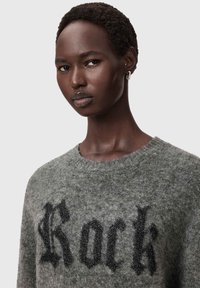 Lähikuva henkilöstä, jolla on lyhyet kiharat hiukset ja joka pukeutunut harmaaseen pörröiseen neuleeseen, jossa on mustilla goottilaisilla kirjaimilla edessä sana "Rock".