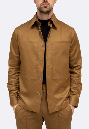 Uomo che indossa una giacca marrone con bottoni sopra una maglietta nera, con le mani nelle tasche dei pantaloni marroni abbinati, davanti a uno sfondo chiaro uniforme.