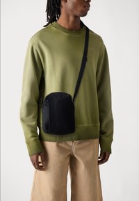 Schwarze Umhängetasche aus robustem Material, mit runder Form, einstellbarem Schultergurt und dezentem Branding. Getragen über einem olivgrünen Sweatshirt.