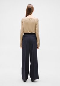 Bézs hosszú ujjú felső mélyen kivágott nyakkivágással, sötétkék, bő szárú nadrággal, gombos díszítéssel és hátsó zsebekkel. Egyszerű háttér.