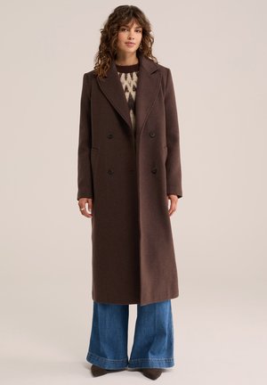 Femme portant un long manteau marron foncé à double boutonnage par-dessus un pull à motifs et un jean bleu à jambes larges, debout devant un fond uni.