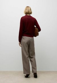 Woman with short blonde hair wearing a burgundy sweater, plaid trousers, and brown high-heeled boots, holding a brown leather bag.