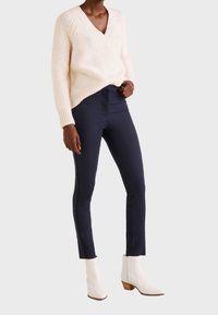Pletený krémový svetr s výstřihem do V, navy slim-fit kalhoty a bílé kotníkové boty na klínovém podpatku; ležérní textura a přiléhavý silueta.