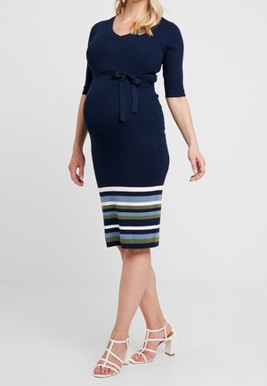 Schwangere Frau in einem eng anliegenden marineblauen Strickkleid mit mehrfarbigen Streifen am Saum und weißen, geschnürten Sandaletten mit Absatz, steht.