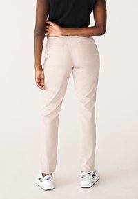 Osoba stojící s levou rukou opřenou o dolní část zad, oblečená v béžových kalhotách, černém topu a bílých teniskách Nike Air.