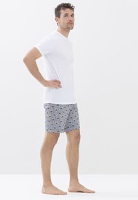 Camiseta blanca de manga corta y pantalones cortos con un diseño estampado en verde y negro sobre un fondo gris a rayas. Descalzo, de pie de lado.