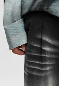 Hand resting beside dark gray herringbone trousers with pleats, wearing light blue textured long-sleeve top against a plain background.