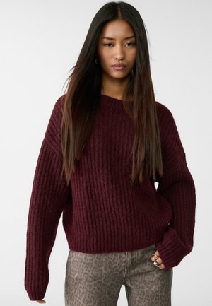 Bordeauxfarbener, gerippter Strickpullover mit weiten Ärmeln und lockerer Passform, kombiniert mit Jeans im Leopardenmuster. Weiche Textur und lässige Silhouette.