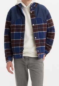 Button-up jacket featuring blue and brown plaid pattern, denim-style shoulders, and snap closures. Paired with a white hooded sweatshirt.