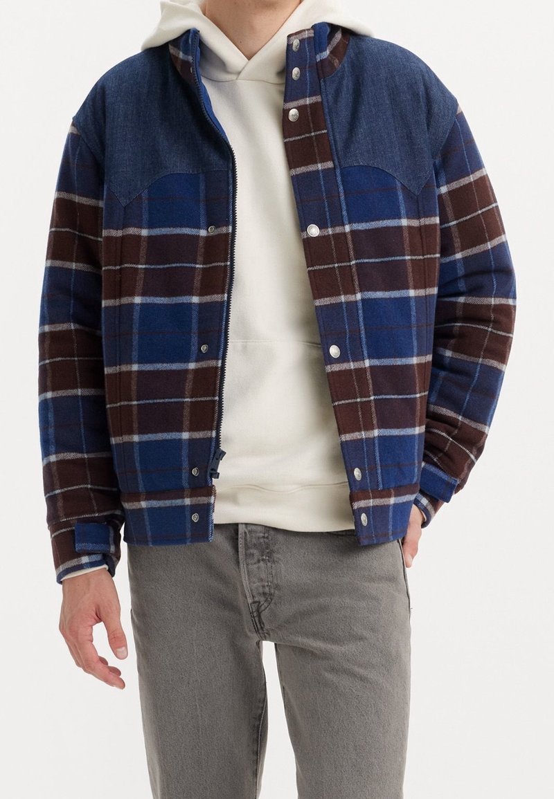 Button-up jacket featuring blue and brown plaid pattern, denim-style shoulders, and snap closures. Paired with a white hooded sweatshirt.