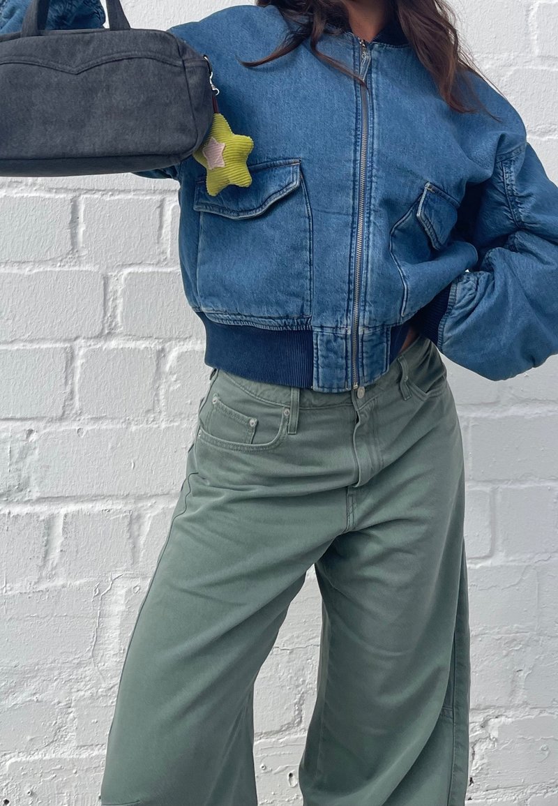 Personne portant une veste en jean bleue courte et un pantalon vert ample, tenant un sac gris avec un porte-clés jaune en forme d'étoile, devant un mur blanc en briques.