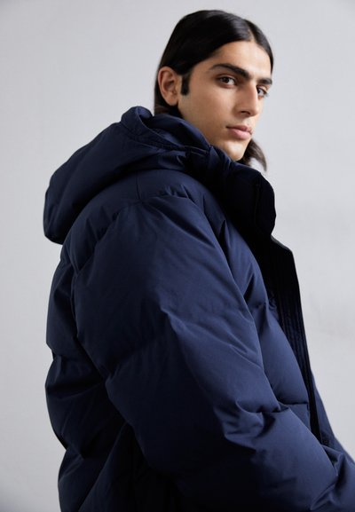 Doudoune bleu marine avec capuche, présentant une surface texturée et un devant zippé. Fabriquée avec un design plus épais et isolé.