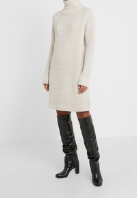 Mujer vistiendo un vestido de punto de canalé beige de manga larga, combinado con botas de cuero negras de tacón alto y caña baja.