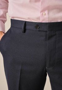 Pantalones de tejido azul marino con frente plano, pasadores para cinturón, cierre de botón y bolsillos laterales, combinados con una camisa de patrón rosa claro.