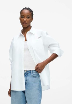 Žena s vrkočmi, ktorá má na sebe nadmerne veľkú bielu košeľu s malým mačacím motívom na vrecku, biele tielko a modré džínsy.