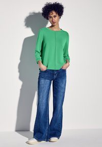 Felpa verde con maniche lunghe e polsini abbinata a jeans a zampa d'elefante blu. L'outfit include sneakers bianche e presenta una superficie liscia.
