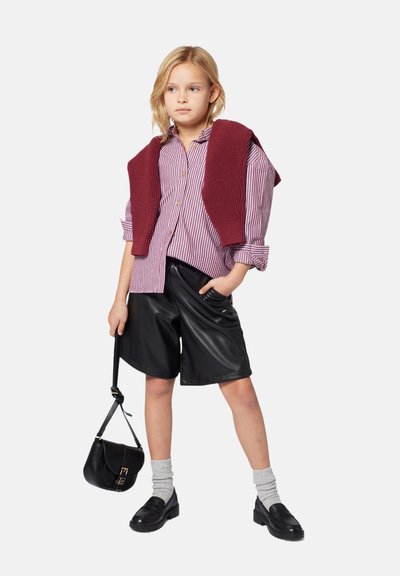 Chemise à rayures rouge et blanche, associée à un short en cuir noir. Pull en maille rouge foncé drapé sur les épaules. Sac et chaussures noirs.