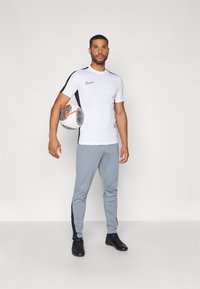 Nike sportoutfit bestaande uit een witte T-shirt met korte mouwen en zwarte zijaccenten, en grijze afgebelde broek met zwarte zijstrepen, met een voetbalsbal vastgehouden.