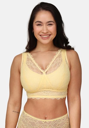 Smiling woman with long dark hair wearing a yellow lace bralette and matching high-waist lace underwear against a plain background.