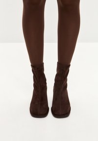 Botine din suede maro cu un design căzut, vârf pătrat și talpă plată. Textură netedă și detalii cusute vizibile.