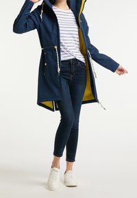 Námořnická voděodolná bunda s kapucí, žlutou podšívkou, stahovacími pasy a bočními kapsami. V kombinaci s tmavými džínami a bílými teniskami.