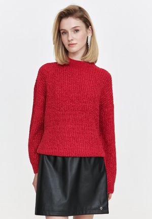 Kobieta z blond włosami, ubrana w czerwoną sweter, czarną skórzaną spódnicę oraz długie wiszące kolczyki, stojąca na tle białej ściany.