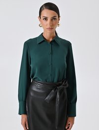 Una donna con i capelli slicked-back indossa orecchini a cerchio dorati, una blusa button-up verde scuro e una gonna di pelle nera legata in vita.