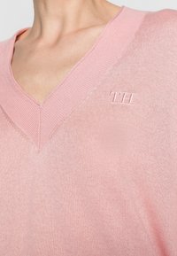 Gros plan sur une personne portant un pull rose à col en V avec un logo "TH" brodé sur le haut de la poitrine.