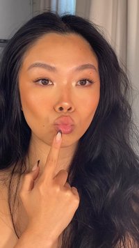 Modell med langt svart bølgete hår, lys hud. Hun lager en kyssende grimase og holder fingeren opp mot leppene. Neglene er lange med en gradient.