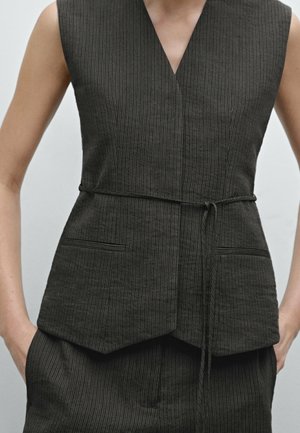 Veste sans manches à fines rayures sombres avec col en V et ceinture fine nouée à la taille, portée avec un pantalon assorti à fines rayures, mains dans les poches.