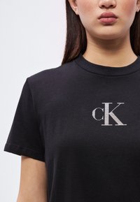Svart bomulls t-skjorte med korte ermer, med en sølvbrodert "cK"-logo på brystet. Jevn tekstur, klassisk rund hals.