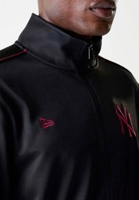 Giacca nera con zip e colletto alto. Presenta cuciture bordeaux e loghi sul petto. Texture liscia con design aderente.