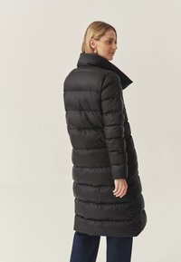 Cappotto lungo imbottito nero con colletto alto, motivo a quadri orizzontali e una texture liscia e lucida. Adatto per il clima più fresco.