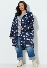 Donna che indossa un impermeabile blu navy a fiori sopra una camicia a righe, jeans risvoltati e stivali bianchi con lacci, in piedi contro uno sfondo bianco.