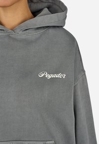 Sivá bavlnená mikina s veľkou prednou kapsou, kapucňou na šnúrku a s nápisom "Pegador" vytlačeným bielym kurzívou na oblasti hrudníka.