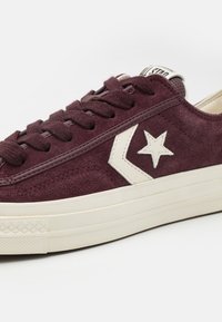 Vinröda mockasneakers med vita gummisulor och snören, med en vit stjärnlogotyp och detaljer längs sidorna. Texturerad yta.