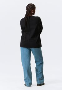 Camiseta negra de manga larga, corte holgado, combinada con pantalones vaqueros de pierna ancha en azul claro. La modelo lleva el cabello trenzado y pendientes de aro plateados.