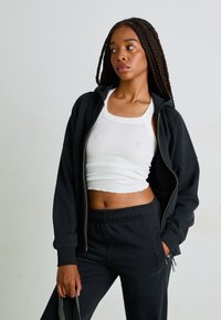 Sweatshirt preto com fecho, por cima de uma blusa de crop top branca com textura canelada, combinado com calças de treino pretas e ajustadas. Cabelo trançado, fundo neutro.