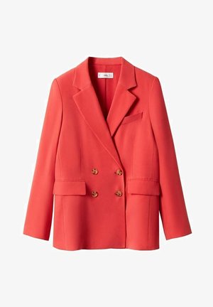 Donna che indossa un blazer doppiopetto corallo, pantaloni su misura abbinati, top bianco e mule aperte con tacco corallo, in piedi su uno sfondo neutro.