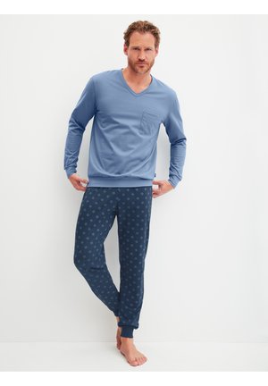 Mann in blauem, langärmligem V-Ausschnitt-Shirt und dunkelblauer, gemusterter Schlafanzughose, barfuß stehend vor weißem Hintergrund.