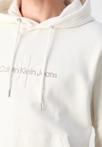 Vit huvtröja i mjukt bomullsblandat tyg. Har snoddar med silverfärgade metalldetaljer och en präglad logotyp för "Calvin Klein Jeans."