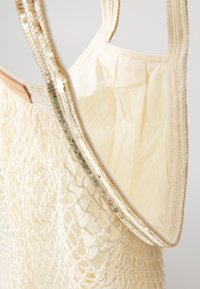 Beżowa torebka szydełkowa o siatkowej teksturze i miękkiej podszewce. Posiada połyskujące, irydyscencyjne akcenty wzdłuż pasków oraz plisowany design.