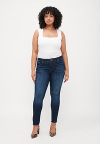 White square-neck tank top paired with dark blue skinny jeans featuring a hem detail. Model wears brown leopard print heels.