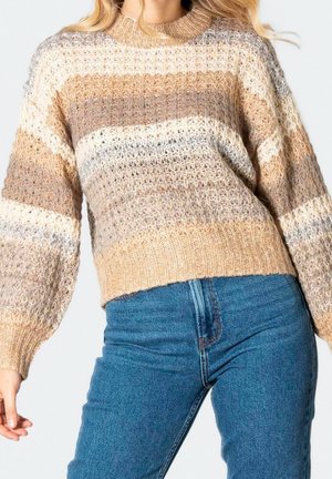 Femme portant un pull en maille rayé beige, marron et crème avec des manches ballon et un jean bleu taille haute sur un fond uni.
