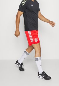Pantaloni rossi del Bayern Monaco con strisce bianche, maglia nera Adidas, calze bianche con strisce grigie e scarpini neri con accenti bianchi.